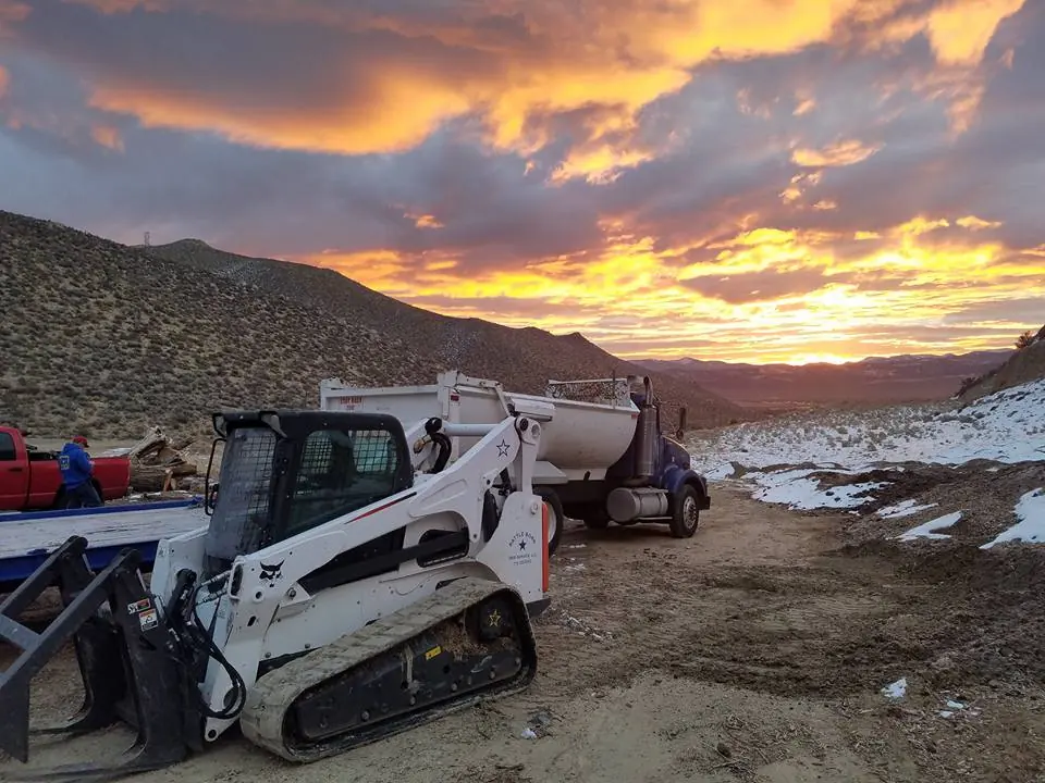 Bobcat track loader and dump truck on a hillside lot at sunset